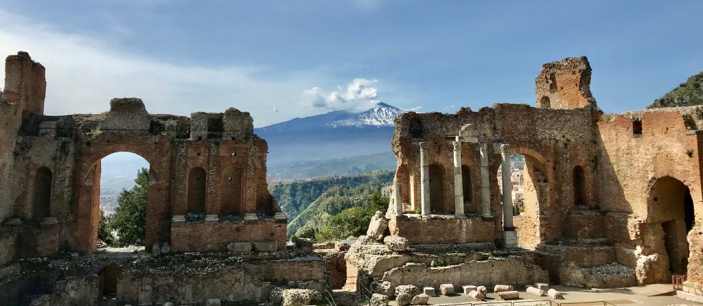 The ancient theater of Taormina in the foreground with Mt. Etna giving off smoke in the distance. 