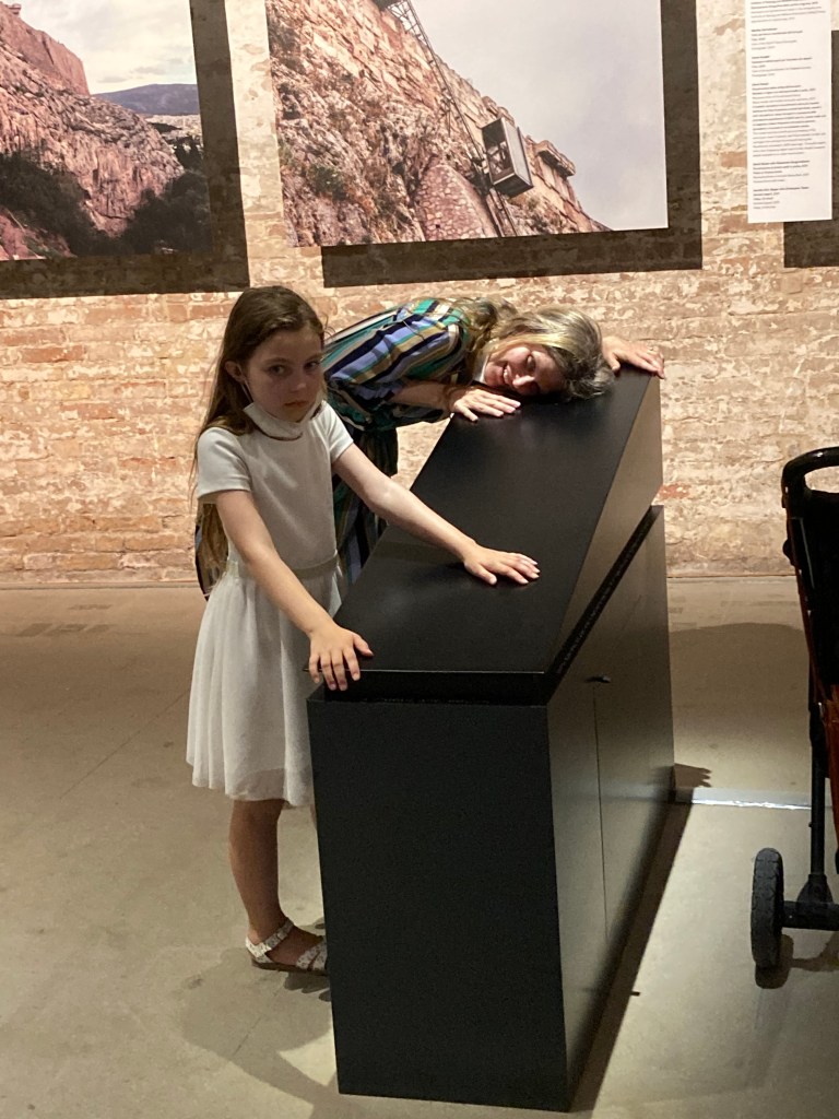 A woman in a striped dress presses her cheek to the surface of a sleek, black model of a ramp, which vibrates gently. In front of her a girl touches the surface of the ramp with her hands and faces the camera. 