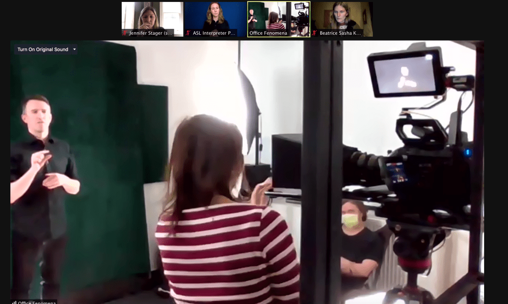 A central screen shows a man wearing a black, button-down shirt standing in front of a deep green background gesturing with his right hand. A woman wearing a face mask and a striped shirt looks at the large movie camera focused on the man. Against the wall another man wearing a face mask and a t-shirt sits against the wall.