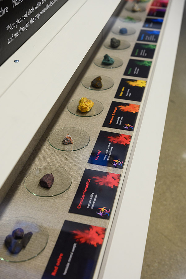 A photo of a line of different pigments, including carbon black, red ochre, cinnabar, red lead, orpiment, and malachite.