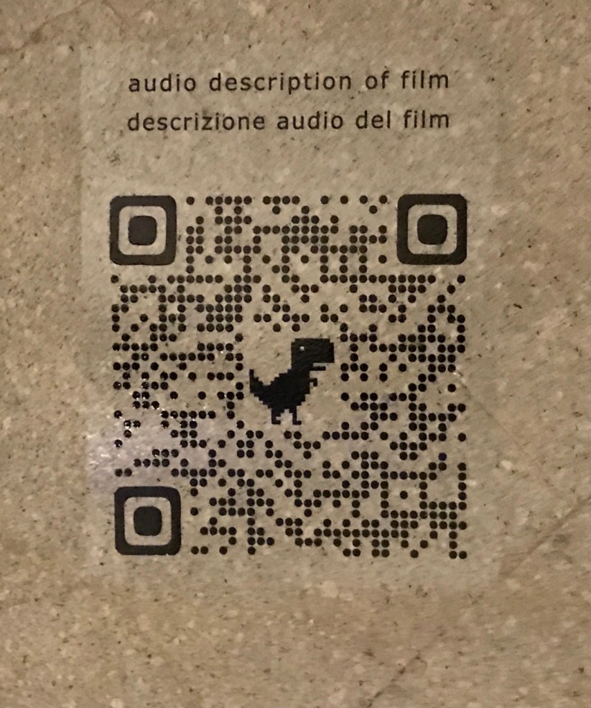 A QR code with a small dinosaur at its center is labeled with Audio Description of Film in English and Italian.