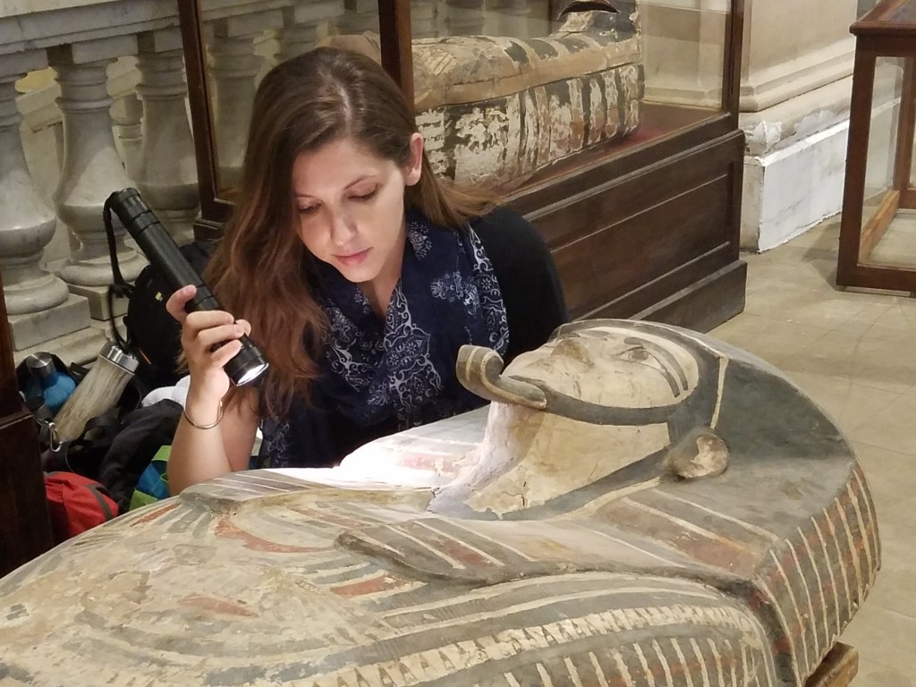 Dr. Danielle Candelora examining an Egyptian mummy coffin with a flashlight in a museum.