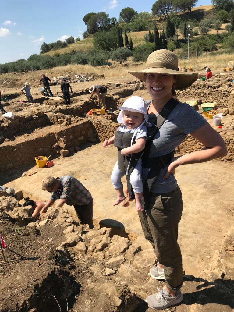 Dr. Katherine Huemoller on excavation standing in a trench with her child strapped to her chest.