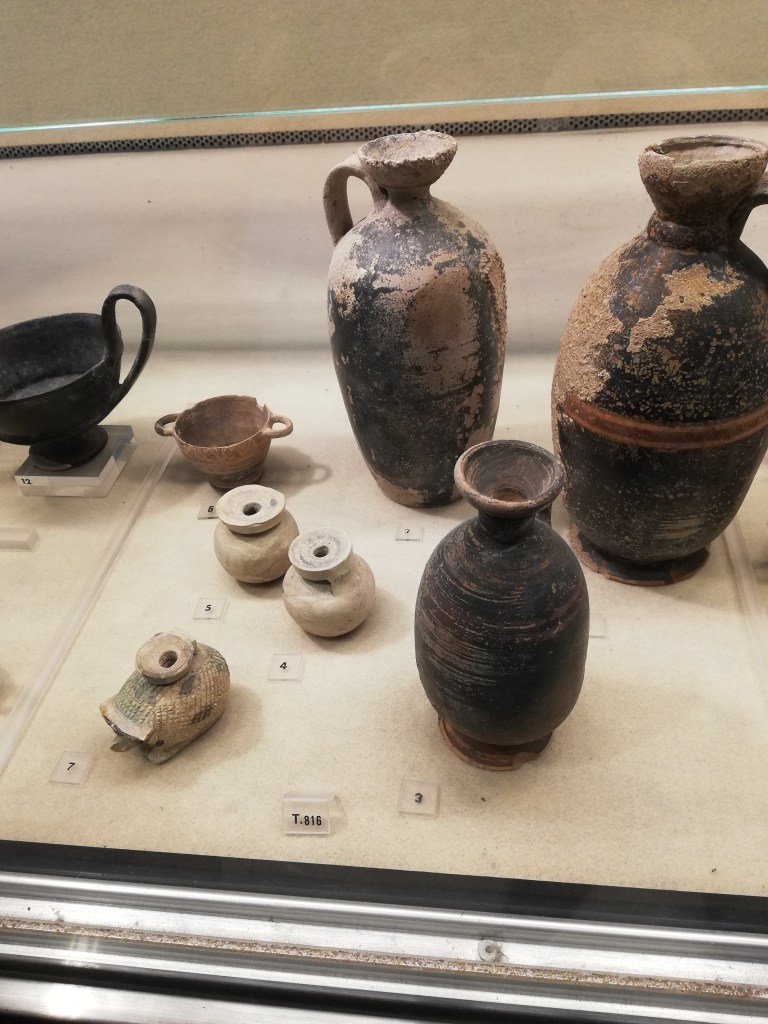 Grave goods in a museum display, include a hedgehog-shaped aryballos and three vases from the Museo Archeologico Regionale, Syracuse.