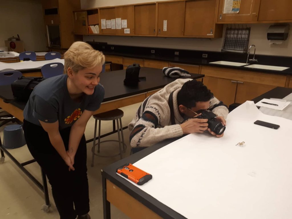 Two students photographing a small object laid out on a white background.