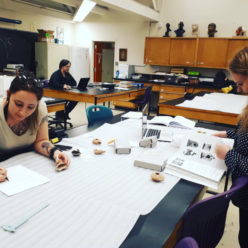 Two students at a table analyzing some lamps. One is writing, the other is looking at a textbook.