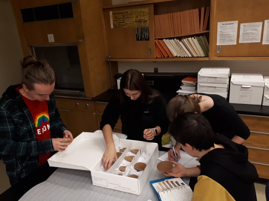 Students gathered around a box of pottery, examining the pieces. 