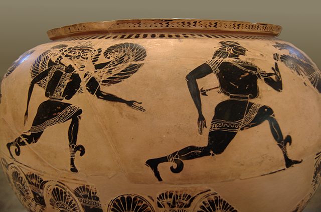 Photograph of a painted terracotta pot. It is a close-up of one side of the pot, where figures painted in black are running. A winged Gorgon with profile legs and arms but frontal face and torso runs after a man with profile face, legs, and head, but frontal torso. Both are wearing tunics and winged sandals. There is decorative black-painted ornaments above and below.