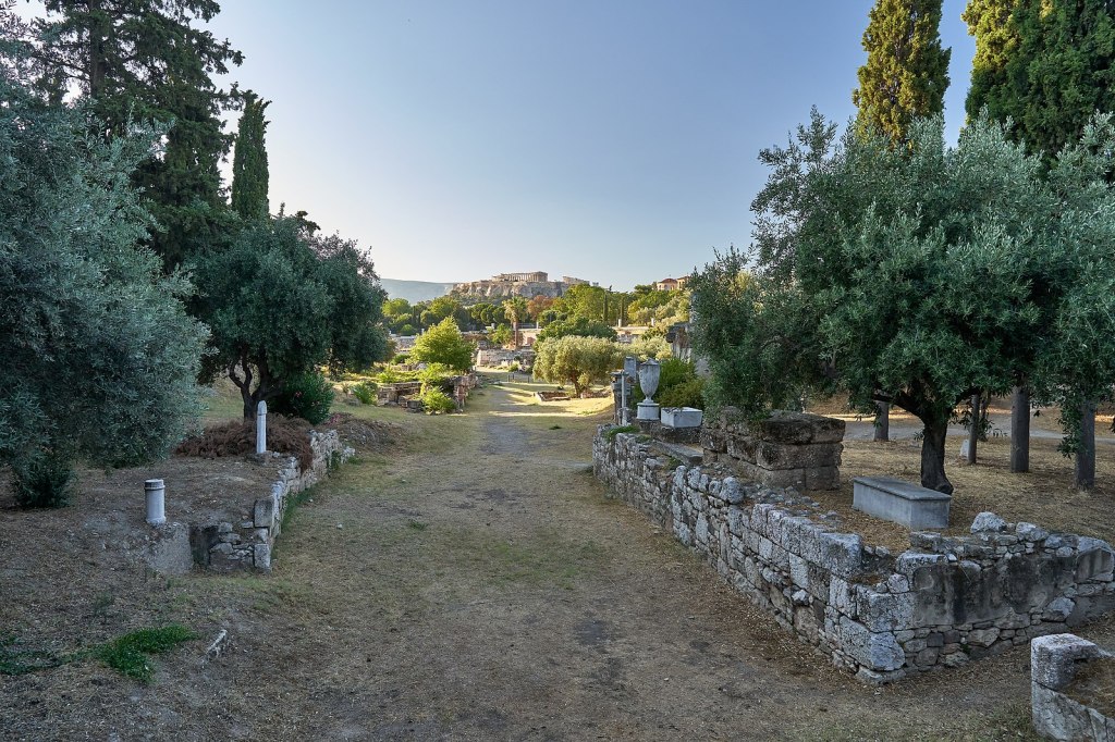 Photo of a grass-grown roadway with stone retaining walls lining either side. Atop these walls are several grave monuments - one looks like a sarcophagus; a few look like columns; one looks like an urn. There are trees on either side of the roadway. In the distance there are more clusters of trees and the Athenian Acropolis.