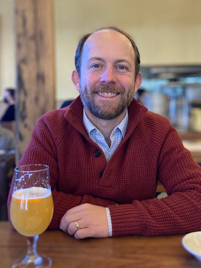 Tate Paulette sits at a table with a glass of beer, smiling at the camera.