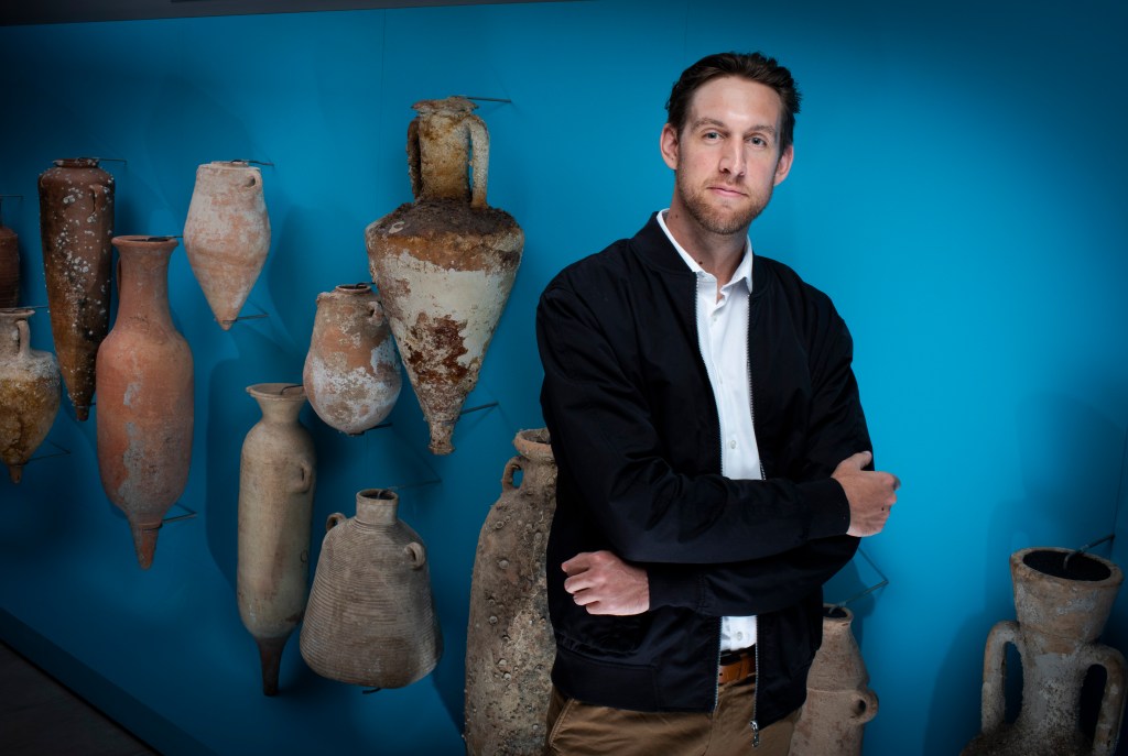 The author stands in front of a bright blue wall with Turkish wine pots of various sizes along it.