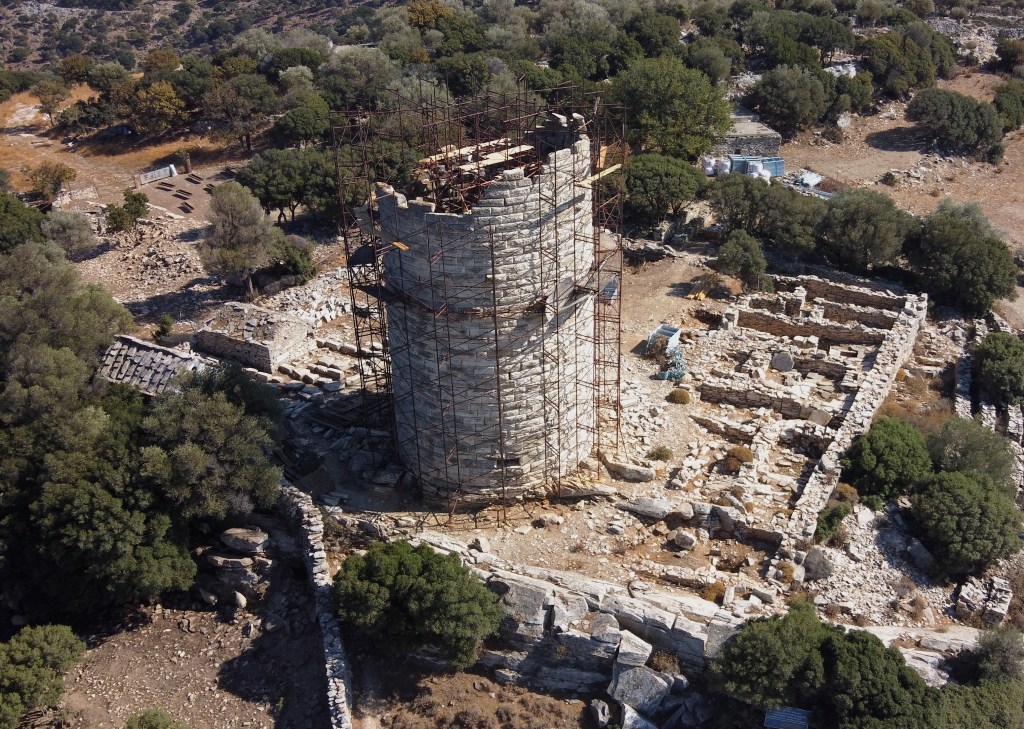 An ancient stone tower surrounded by metal scaffolding stands among ruins and trees