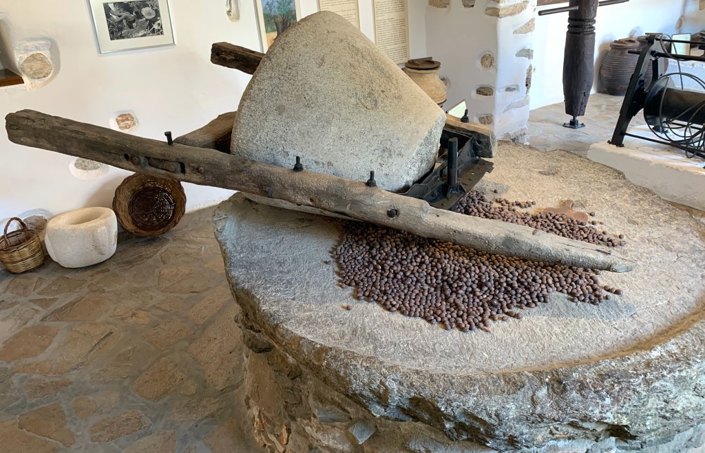 A reconstructed olive press is displayed in a room with woven baskets and other tools. The press is made of stone and metal.