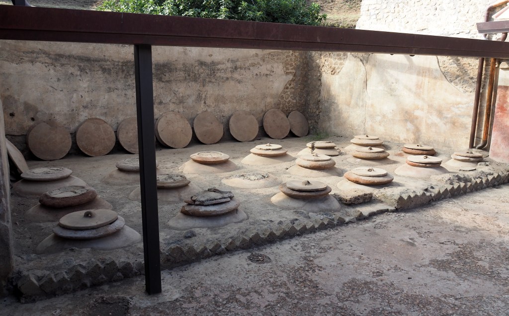 A large stone wine cellar in a stone walled open-air room. The floor has 18 openings with stone lids on them.