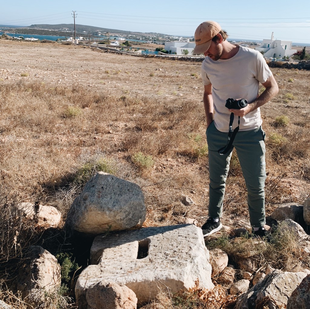 The author stands in a dry grassy field with a camera in his hands, looking at two stone wine press counterweights on the ground.