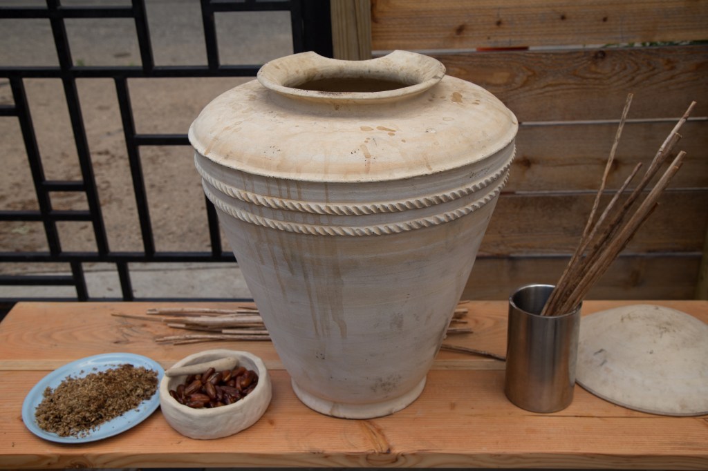 A vessel sits atop a wooden table along with some of the ingredients used such as dates, and a metal cup with reed straws in it