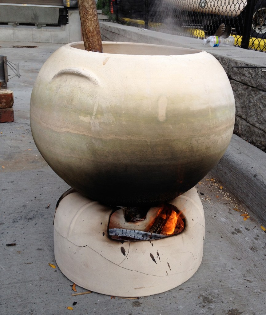 A large round ceramic vessel with a stirring stick inside sits atop a base containing burning wood. Steam is coming out of the top of the vessel.