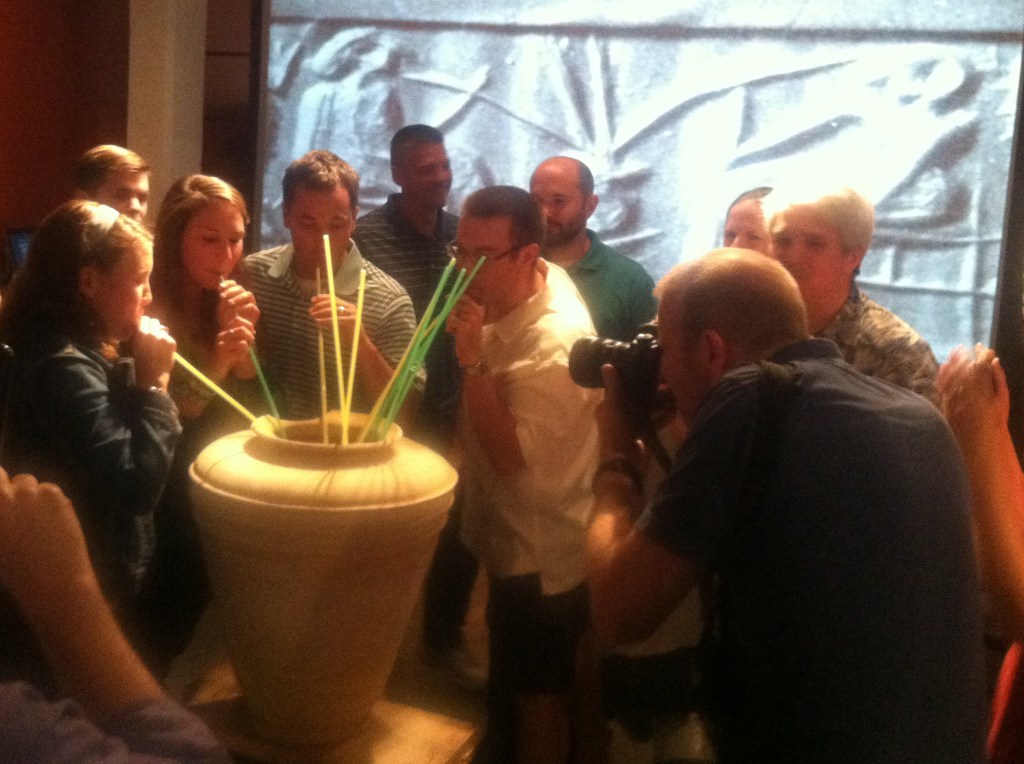 A group of people stand around a vessel filled with beer, drinking from it with long reed straws