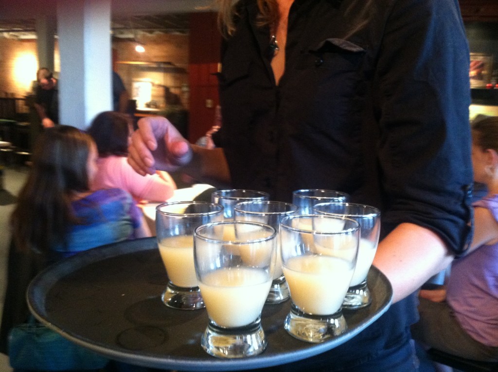 A server holds a black tray with seven glasses of homemade beer on it
