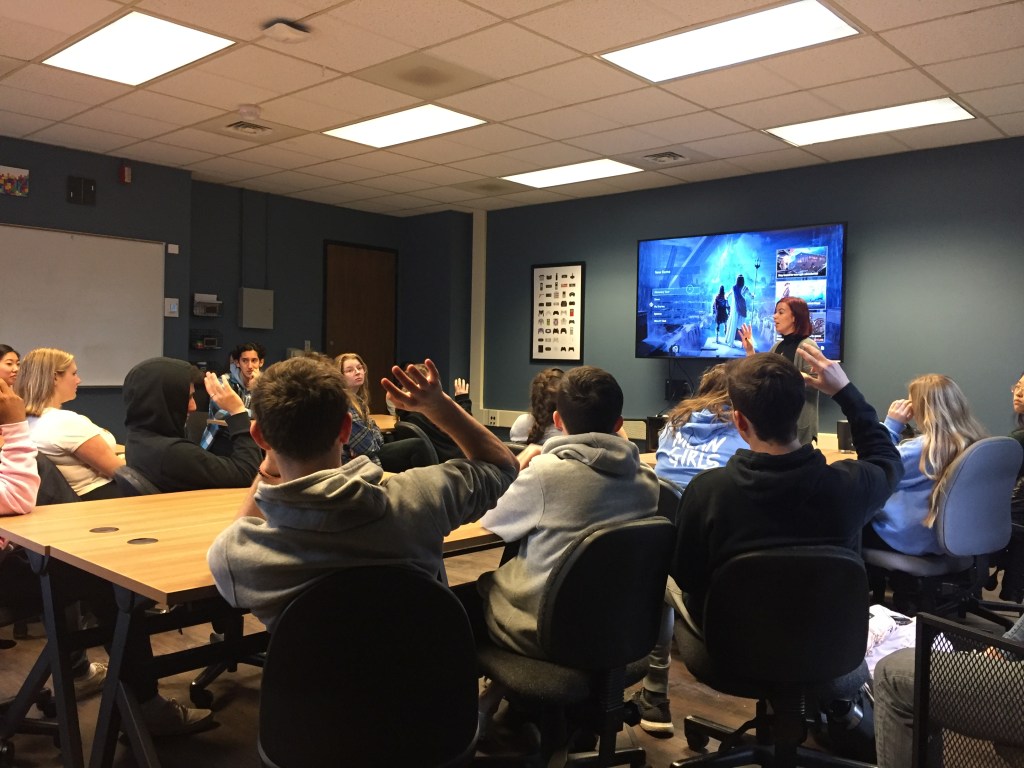 Tine Rassalle stands at the front of a classroom full of students with the Assassin's Creed video game on the screen