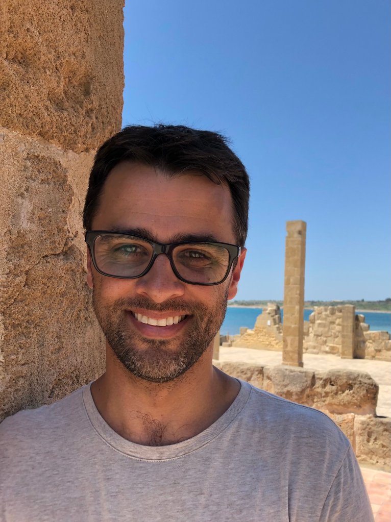 Justin Leidwanger smiles at the camera and is wearing a grey shirt. he is standing beside a stone building. the sky is clear and blue.