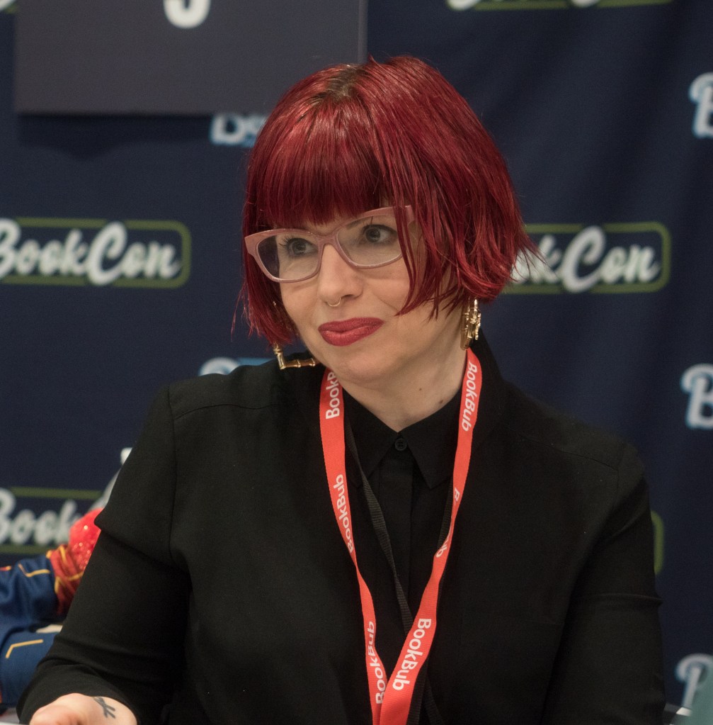 Kelly Sue DeConnick softly smiles. She is wearing a black long sleeved blouse, light red framed glasses, a red lanyard around her neck, and she has short dyed red hair with bangs.