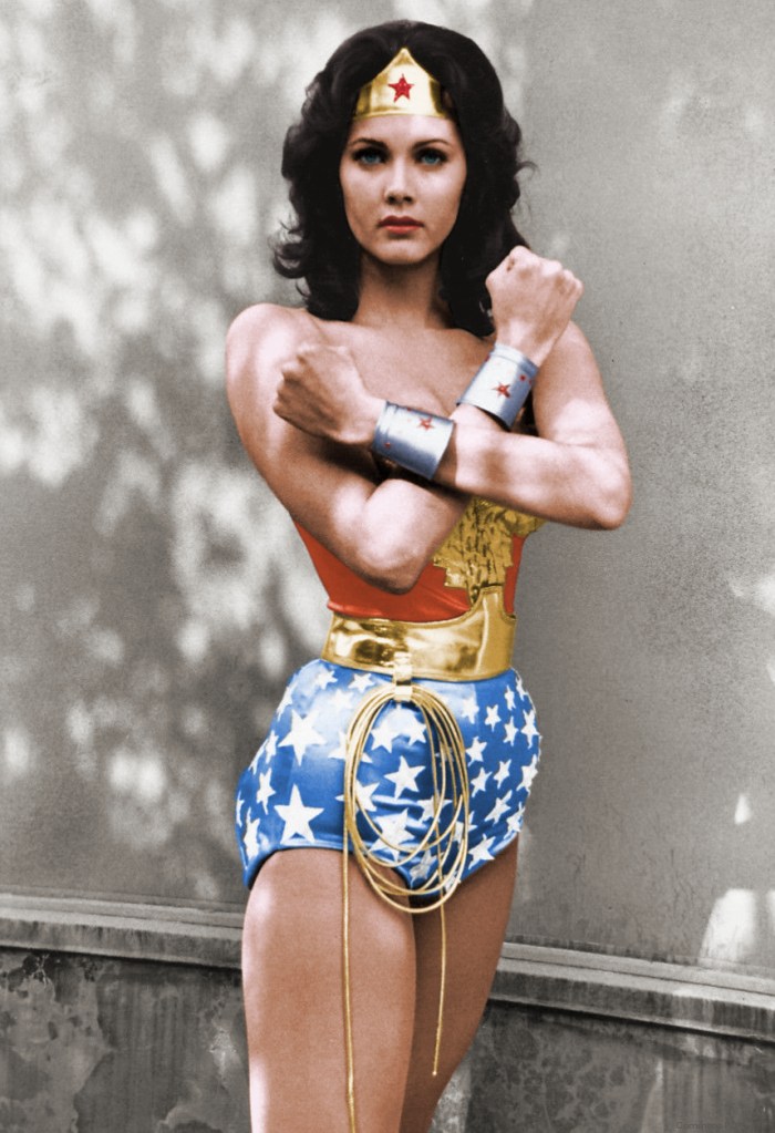 Lynda Carter is dressed in her Wonder Woman costume. She is wearing short blue shorts with white stars, a golden whip on her waist, a golden belt, a small red top, silver cuffs on her wrists, and a golden tiara with a red star in the middle. Her forearms are crossed over her chest in an X formation.