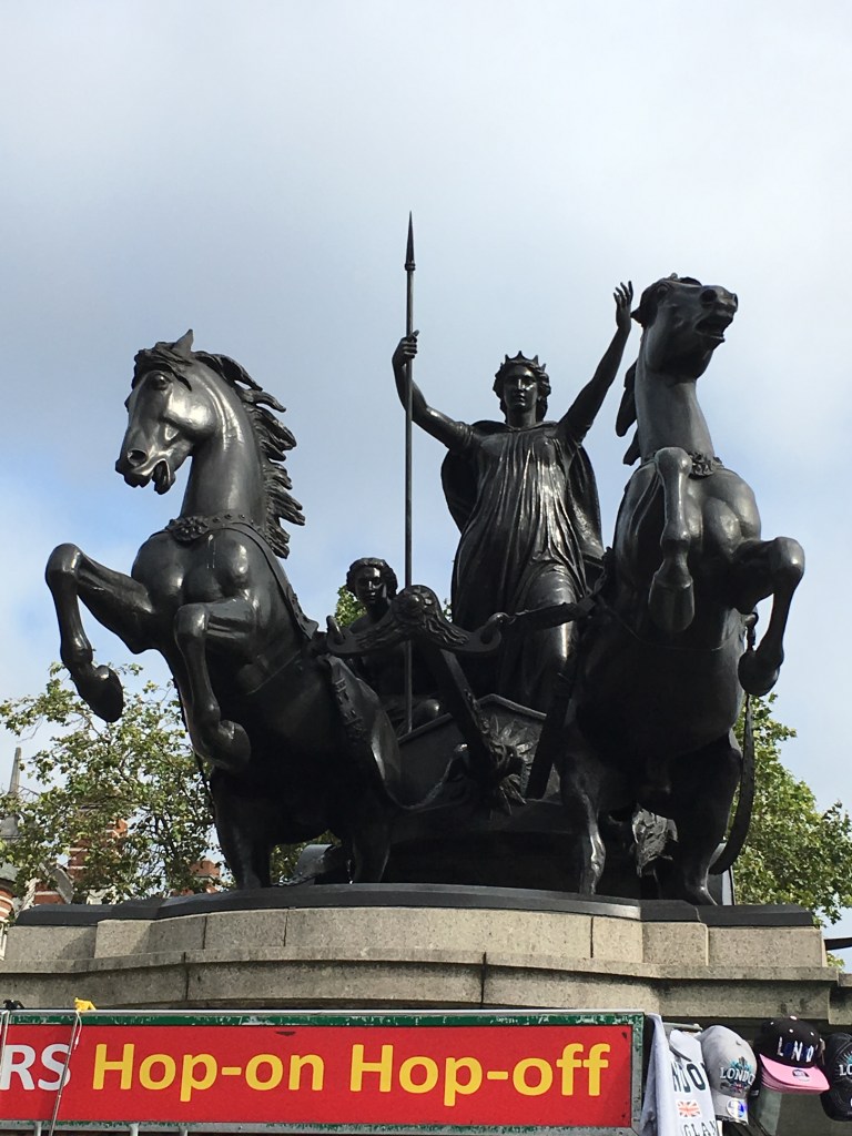 A statue of Boadicea in a chariot pulled by two rearing horses sits atop a building. She stands with her arms raised and a spear in her right hand.