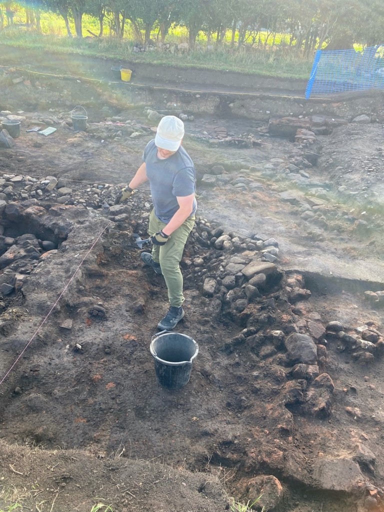 Neil stands on rocky ground within an archaeological excavation. He is wearing a light coloured baseball cap, a blue t-shirt, and green pants.