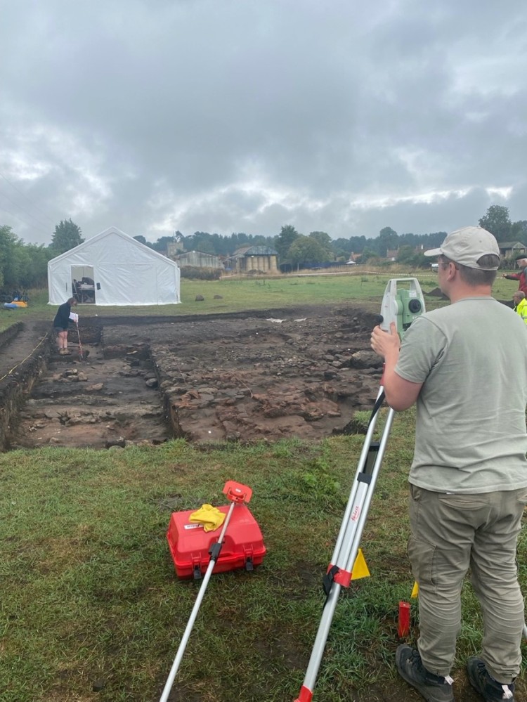 an excavation square has been dug in a large green field, and one person in inside the perimeter examining something. Another person stands in the foreground with survey equipment. 