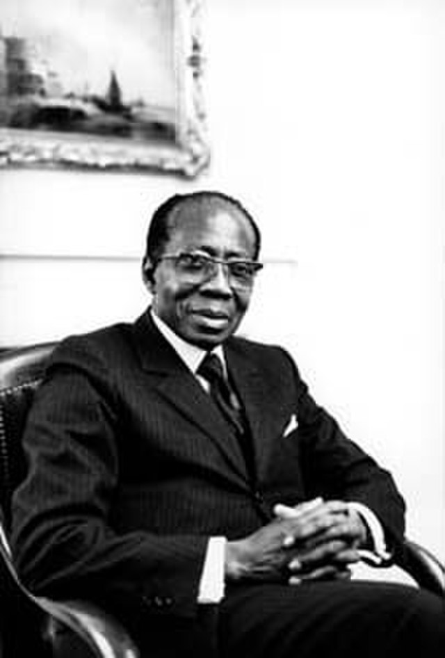 Leopold Senghor sits in a chair with his hands in his lap. He wears a suit with a black jacket and black tie. He has dark skin, and dark short hair. He wears glasses. It is an old black and white photo.