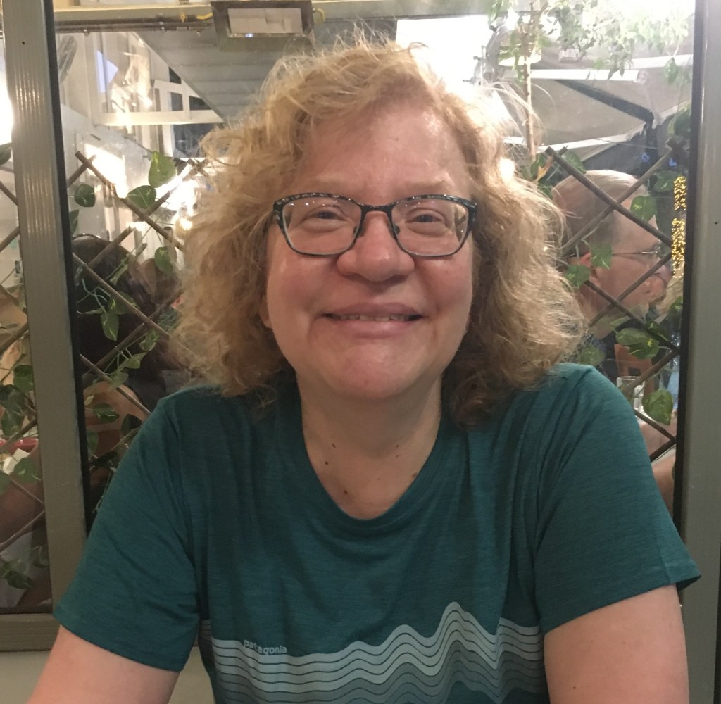 Sandra Garvie-Lok smiles at the camera. She has blonde curly hair, dark rimmed glasses, and a green t-shirt