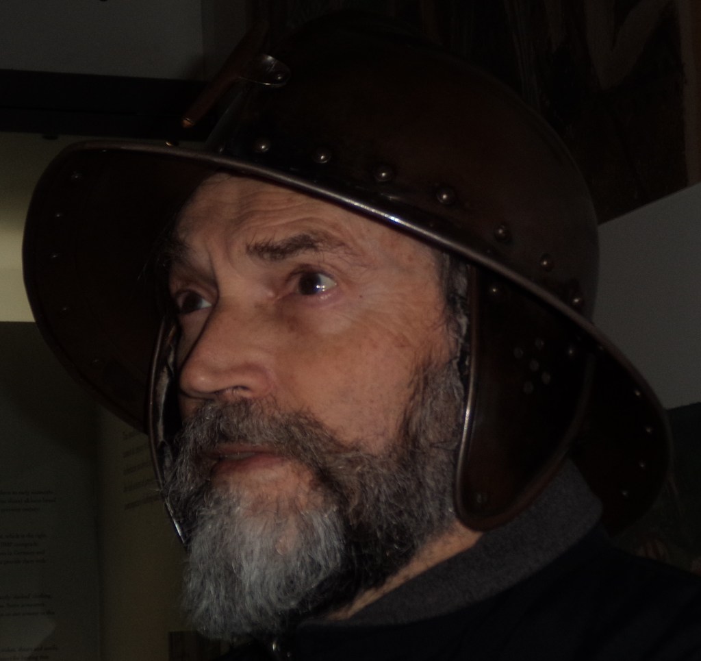 Hector Williams looks to the side. He wears the helmet of a man at arms from the Battle of Agincourt 1415. He has dark eyes, and a black and grey moustache and beard.