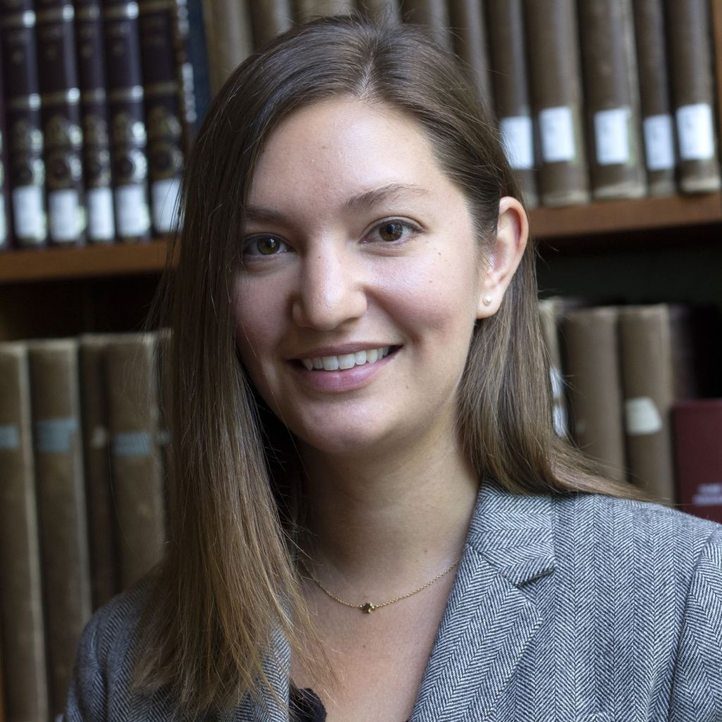 Melissa Cradic smiles at the camera. She has shoulder-length brown hair, with the left side tucked behind her ear. She has brown eyes, and light skin. She is wearing a grey blazer, and has a small necklace on. 