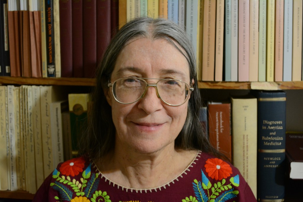 JoAnn Scurlock smiles at the camera. She has long, straight grey and black hair, light skin, and glasses. She wears a floral print blouse with colours of maroon, red, green, blue, and yellow.