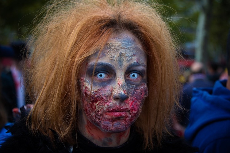 A person dressed as a zombie stares into the camera with white-blue contact lenses. They have short, tousled ginger coloured hair, grey face-paint, and blood coloured makeup smeared around their mouth. 