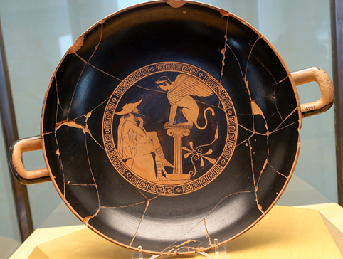 A red figure kylix showing Oedipus sitting down, gazing thoughtfully up at a Theban Sphinx sitting on a pillar.