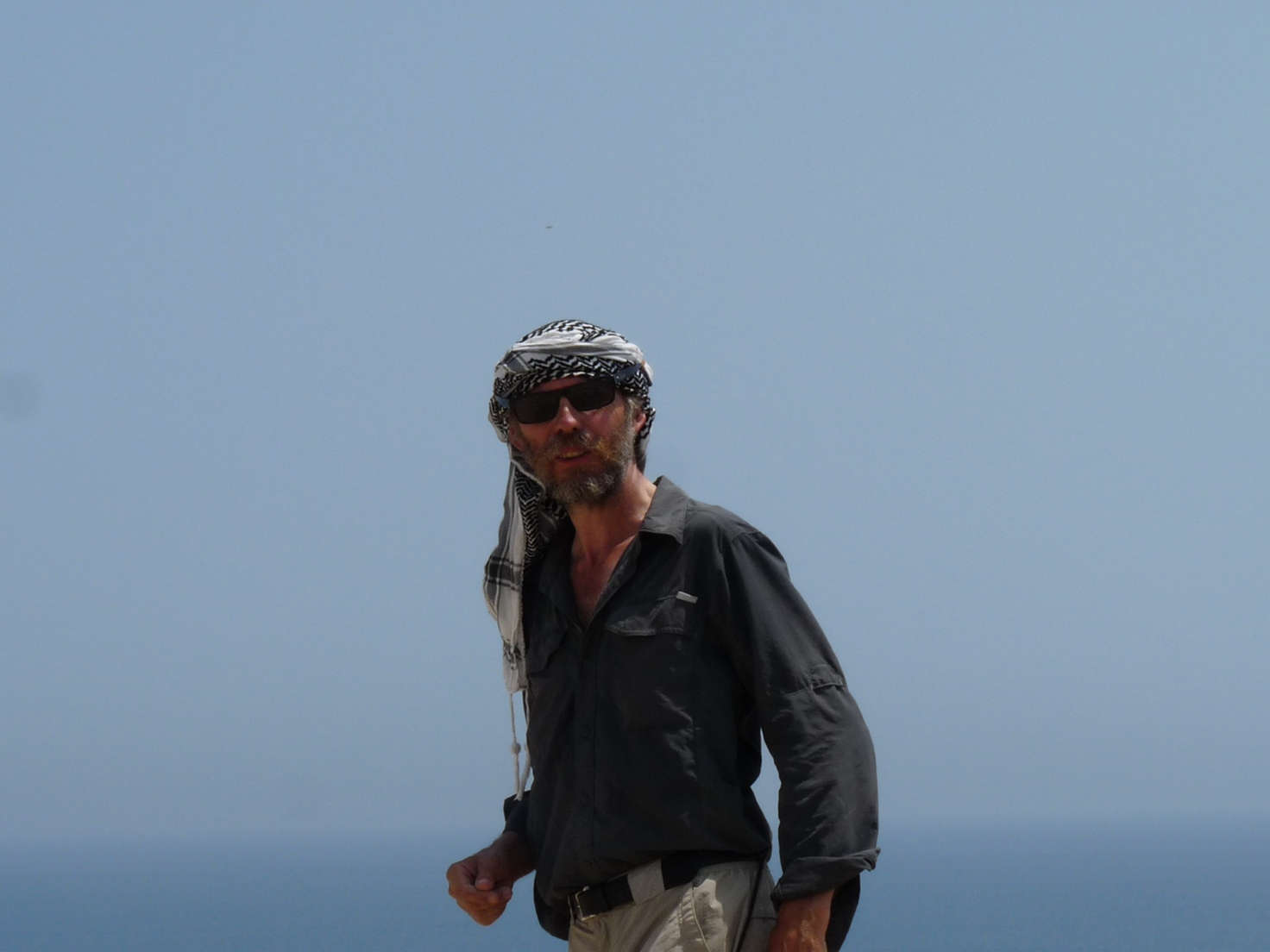 Thierry Petit wears a long grey loose-fitting button up shirt, light grey pants, and a black belt. He has a headscarf on, black sunglasses, and has grey and black facial hair. The background is light blue, and he is presumably somewhere arid.