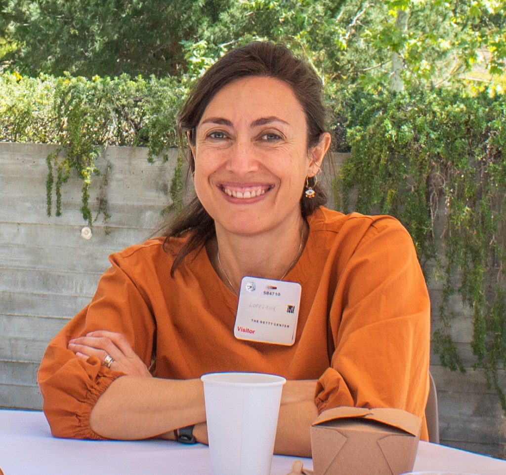 Carolina López-Ruiz sits at a table outdoors on a a sunny day and smiles at the camera with her arms crossed. She has brown hair in a ponytail, and orange blouse.