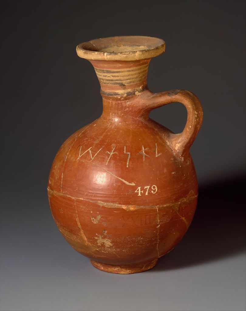 A brown terracotta jug sits on a grey table. It has a rounded body, a single handle, a short neck, and a fluted mouth. It has some Phoenician script carved into it.