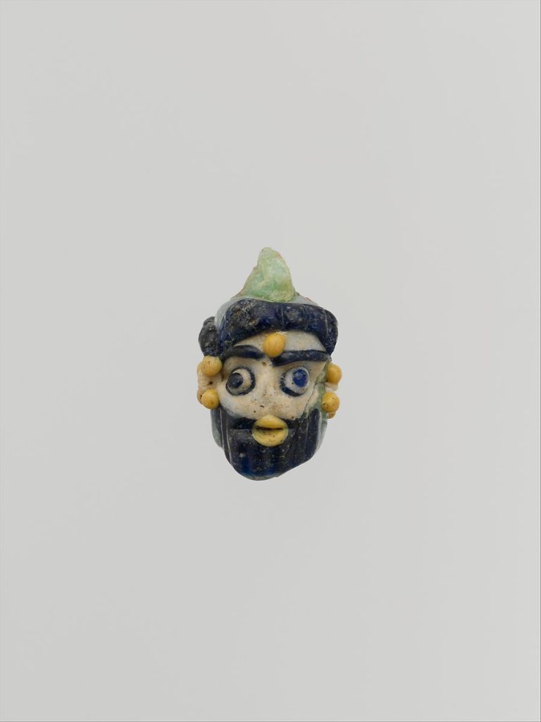 a small glass pendant depicts a man with dark hair, eyebrows, and a beard. He has a green pointed hat, golden beads on his ears, and one in the middle of his forehead.