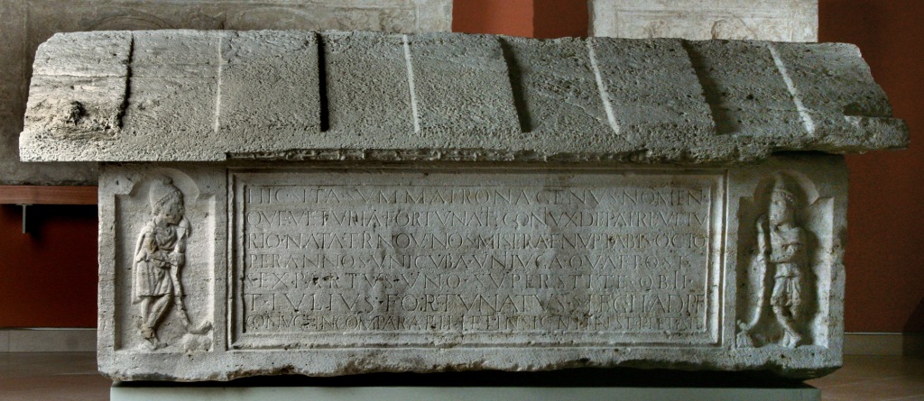 A stone sarcophagus with a gabled roof. There is a long inscription in the centre, and two figures leaning on staffs at either end.