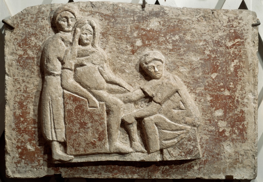 A stone relief of a midwife assisting a birthing woman sitting on a chair. A woman stands behind the pregnant woman, wrapping her in an embrace. 