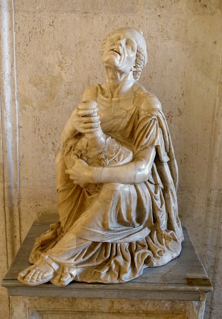 Marble statue of an old woman clutching an amphora of wine in her hands, as she sits on the ground.