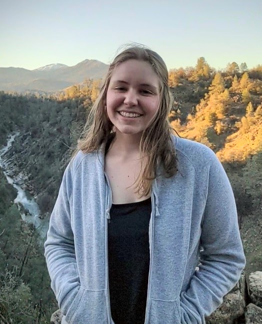 A person standing with black shert and grey zip-up with medium-length blonde hair smiling; a valley with a river can be seen behind. It looks like sunset.