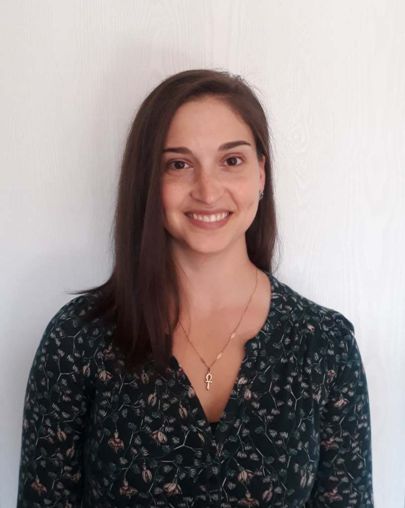 A photos of Roxanne Bélanger Sarrazin, a white woman with medium length, dark brown hair. She wears a floral long sleeve top and stands against a white background. 