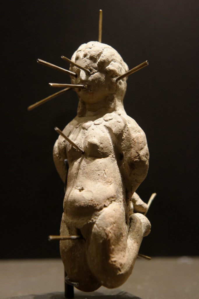 A terracotta figurine of a kneeling woman with several pins sticking through her face, breasts, and pubic region.