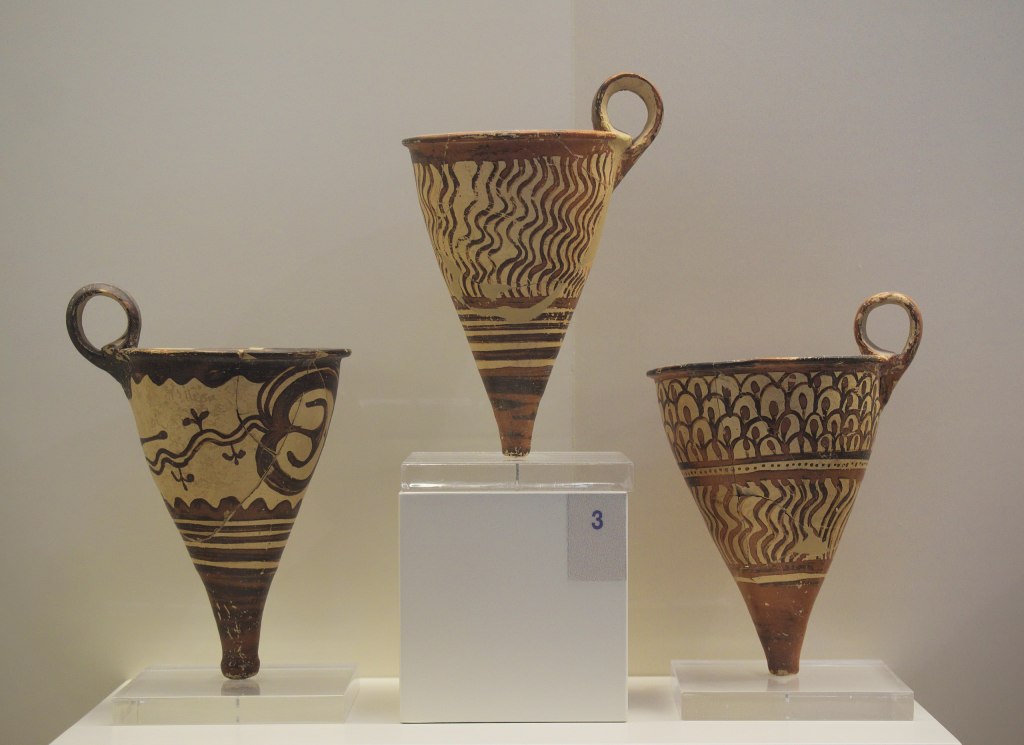 Museum display of three conical cups (rhyta) side by side, with the middle one on a pedestal. They are made of clay and fully decorated in brownish paint (straight lines, solid areas, wavy lines, and other abstract patterns)