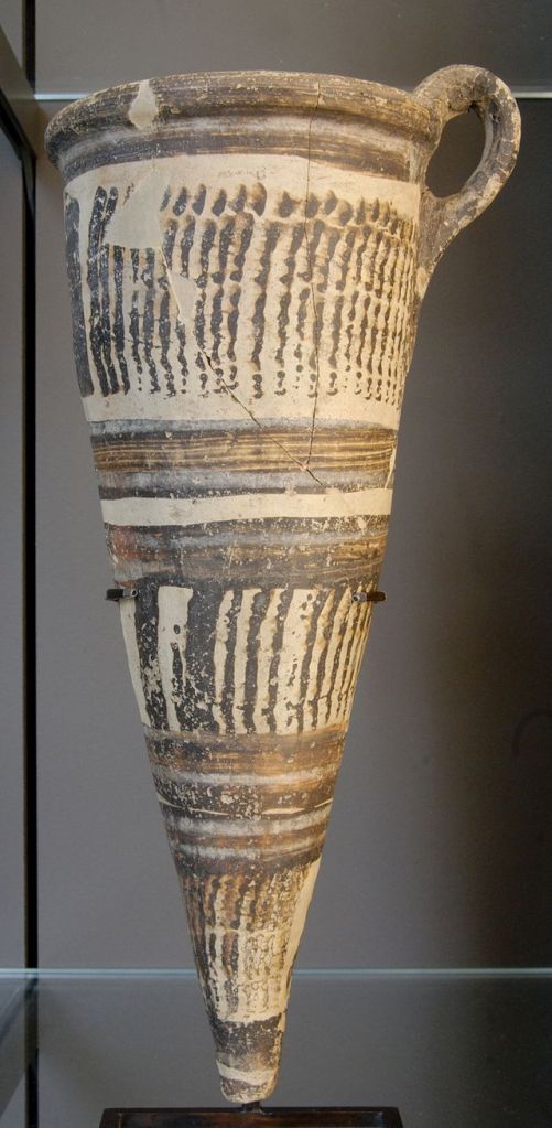 A museum display of a large conical drinking cup (rhyton) with a handle at the top on the right. Decorated in brown glaze (alternating vertical and horizontal lines)