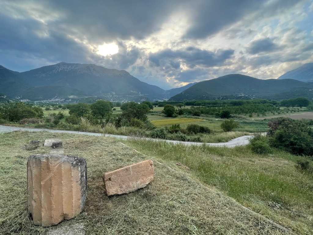 There are broken limestone column drums in the foreground on a small hill. A path runs through bushes and grass behind this mount and there are more fields, bushes, and trees in the background. In the far background are mountains with a cloudy sky overhead and the sun barely breaking through in the late afternoon.